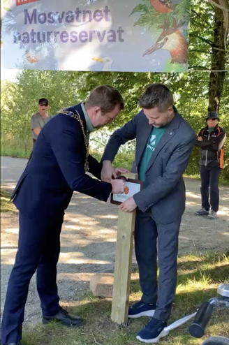 Ordfører Tormod Losnedal og utvalgsleder i klima og natur, Kjartan Alexander Lunde, skrur fast et skilt til en trestolpe ved Mosvatnet.