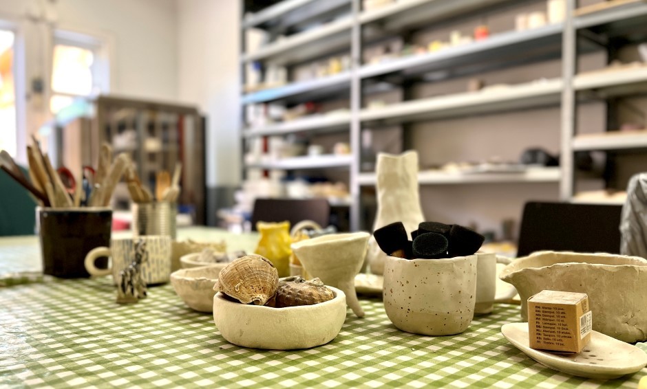 Vi ser bordet inne i keramikkverkstedet på Bergeland bydelshus, med flere små skåler og utstyr laget på verkstedet. Bak bordet ser vi hyller med utstyr.