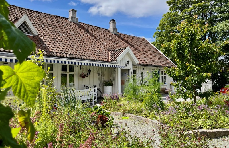 Vi ser det hvite gamle trebygget fra utsiden. Foran ligger en frodig hage i sommersolen.