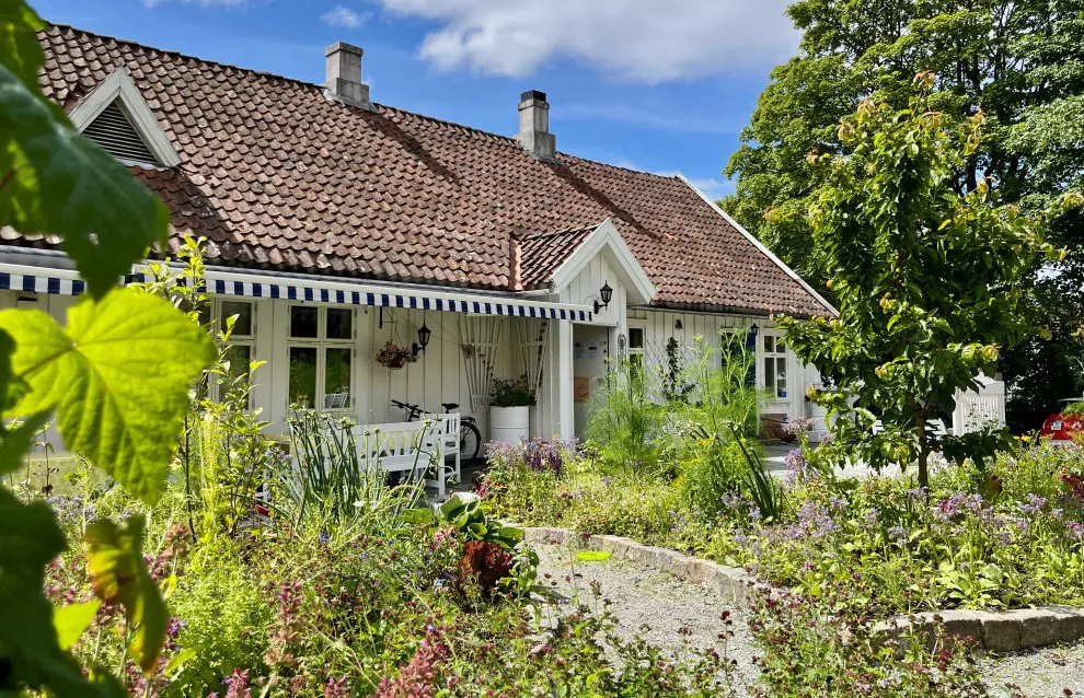 Vi ser det hvite gamle trebygget fra utsiden. Foran ligger en frodig hage i sommersolen.