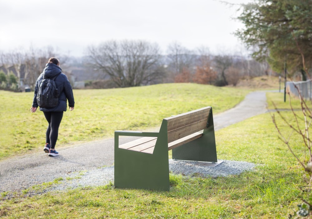 Vi ser et bilde av en benk som står i et grøntområde i Stavanger. En dame går forbi i bakgrunnen. 