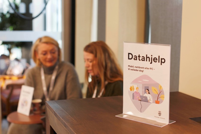 Bildet viser et skilt som det står datahjelp på. Det står på en vindusbenk, og i bakgrunnen ser vi at Tone Hafnor og Elin Halvorsen sitter.