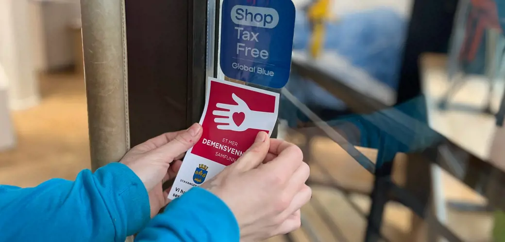 Et godt synlig klistremerke etter fullført kurs gir besøkende informasjon om at dette stedet har kunnskap om demens. To hender som plasserer demensvennlig klistremerket på en inngangsdør.