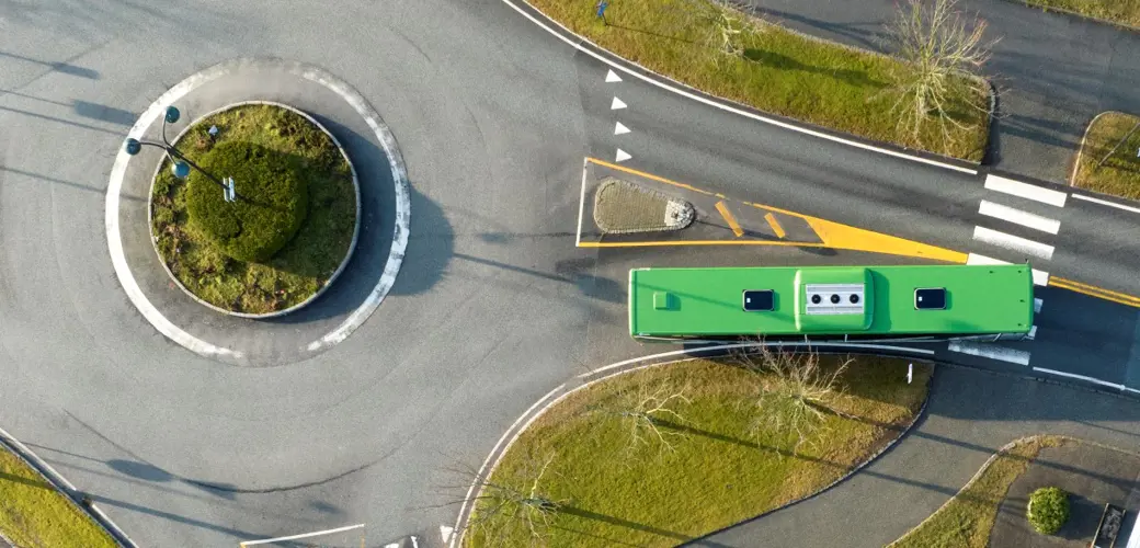 Bildet viser en vei fra luften. Vi ser veistrukturen, rundkjøring, grønne plen, busker og en en grønn buss som kjører på veien.
