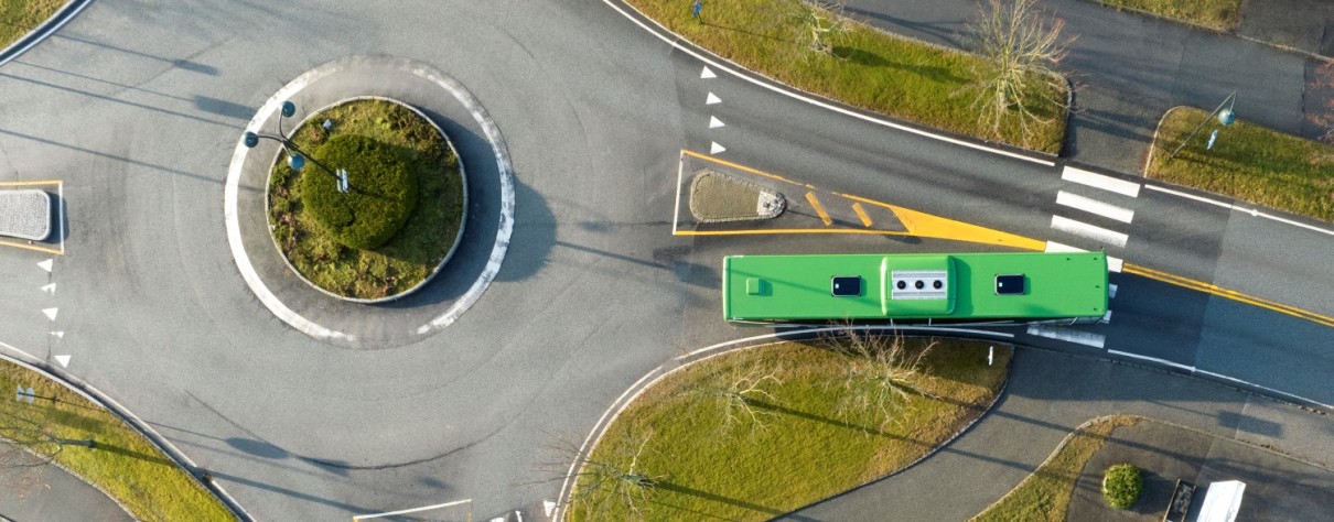 Bildet viser en vei fra luften. Vi ser veistrukturen, rundkjøring, grønne plen, busker og en en grønn buss som kjører på veien. 