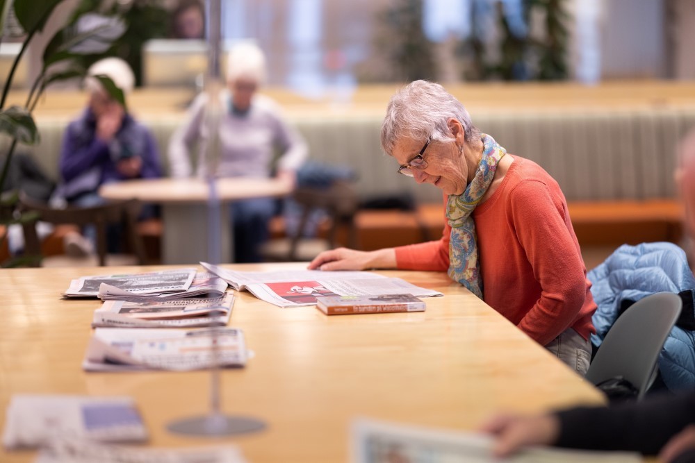 Vi ser en eldre dame som leser en avis. Hun sitter ved det store avisbordet på Sølvberget. I bakgrunnen ser vi andre som sitter.