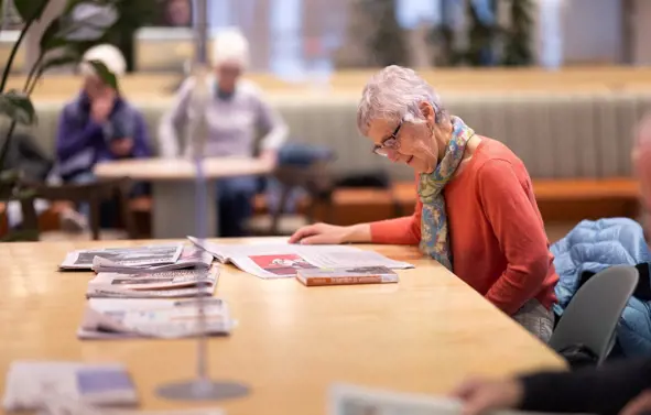 Vi ser en eldre dame som leser en avis. Hun sitter ved det store avisbordet på Sølvberget. I bakgrunnen ser vi andre som sitter.