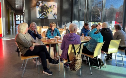 Vi ser åtte damer som sitter rundt bordet ved kafeen i Storhaughallen. 