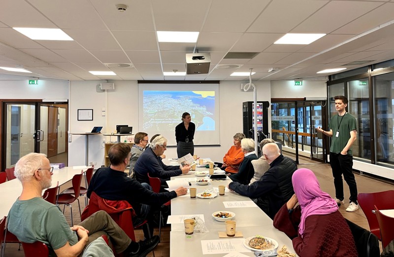 Bildet viser deltakerne som sitter rundt et stort bord og hører på presentasjon av plan for visuell utforming, som blir lagt frem av Stian Alvestad som står foran. 