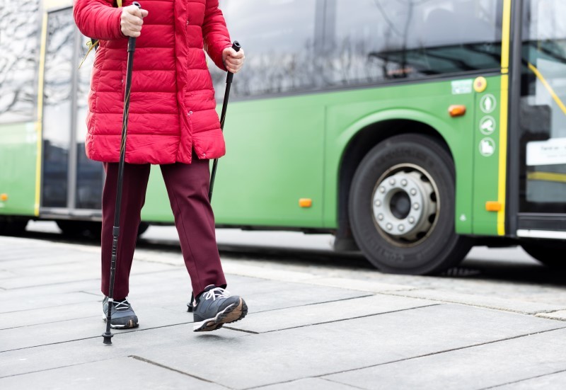 Vi ser en eldre kvinne i rød boblekåpe med gåstaver, som går i byen. Bak henne står en grønn buss fra Kolumbus. 