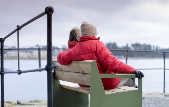 Vi ser en mann og dame som sitter tett sammen på en grønn benk og ser utover sjøen. 