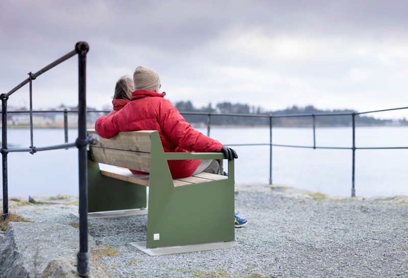 Vi ser et eldre par som sitter på en grønn benk ved sjøen.