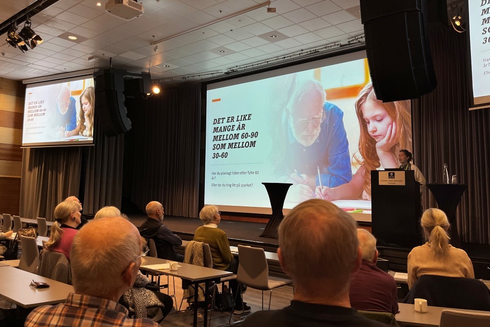 Bildet viser salen på konferansen, med mange mennesker som sitter og følger med på scenen. Der holder Ruth -Karin Haslerud foredrag. På skjermen på scenen står det, "Det er like mange år mellom 60-90 som mellom 30-60".