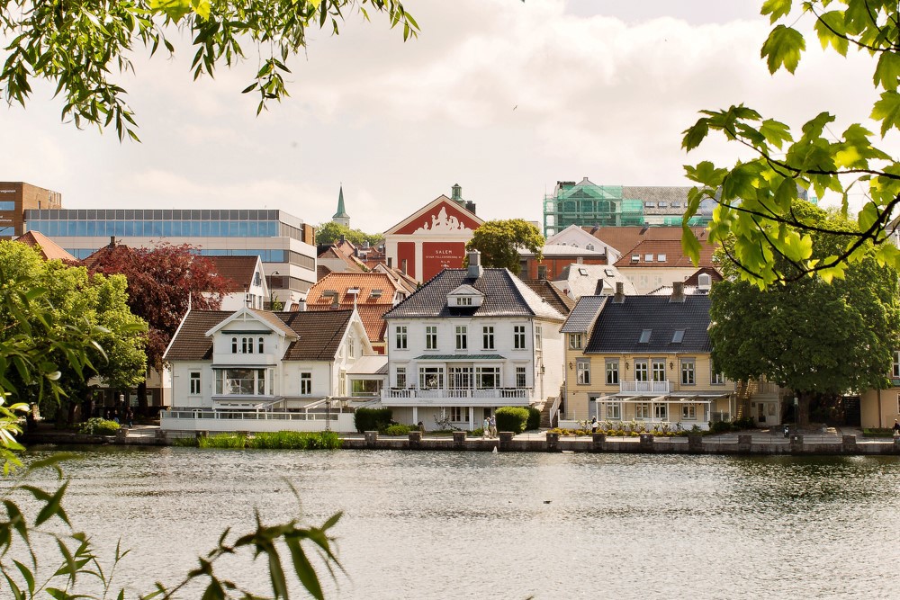 Bildet viser et utsnitt av utsikten ved Breiavatnet. Vi ser flere hus som ligger ved vannet. Det er sommer, grønt og folk går forbi.