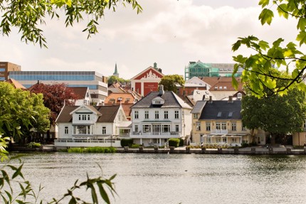 Bildet viser et utsnitt av utsikten ved Breiavatnet. Vi ser flere hus som ligger ved vannet. Det er sommer, grønt og folk går forbi.