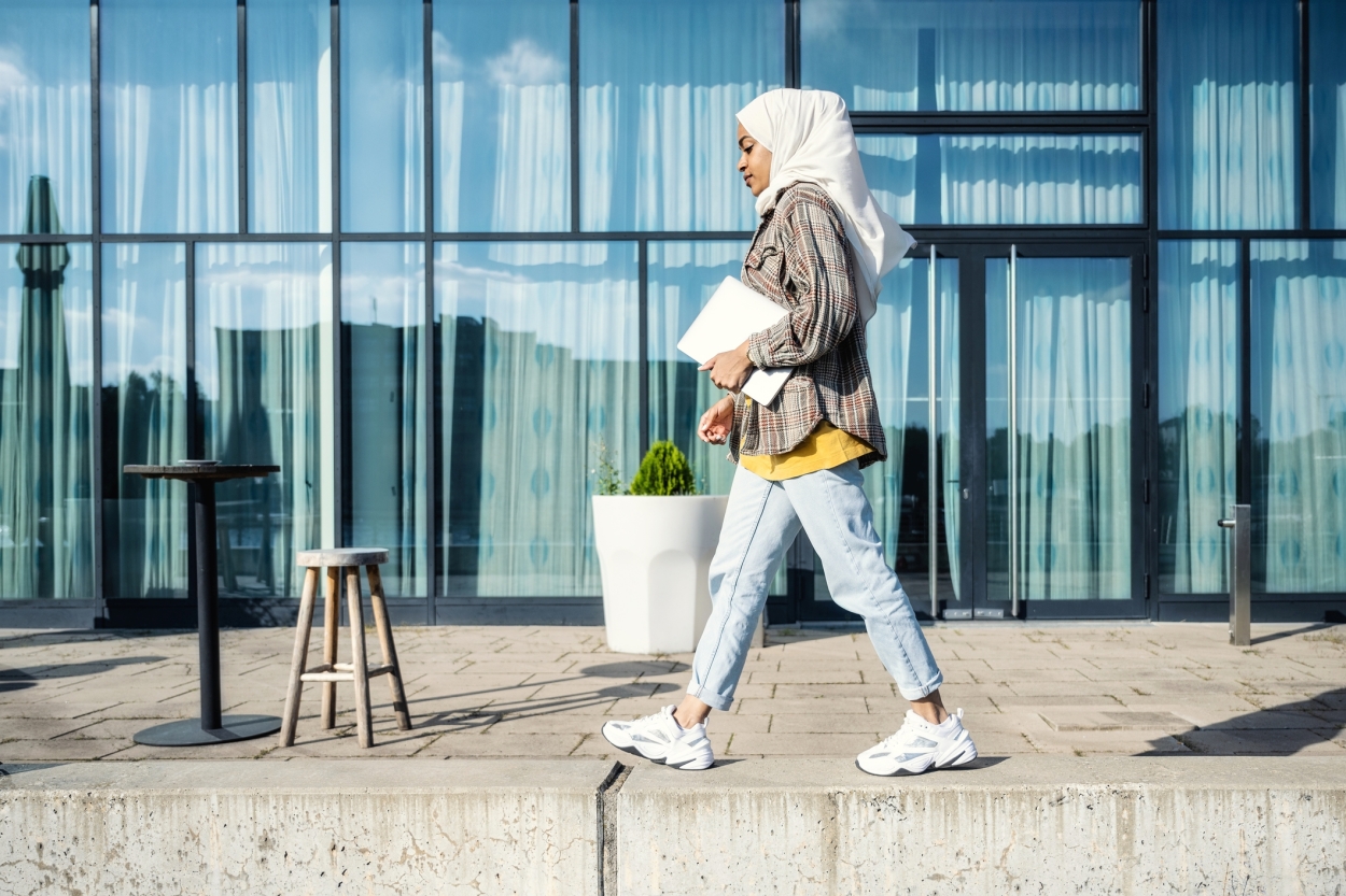 Jente med hijab som går forbi bygning med PC under armen