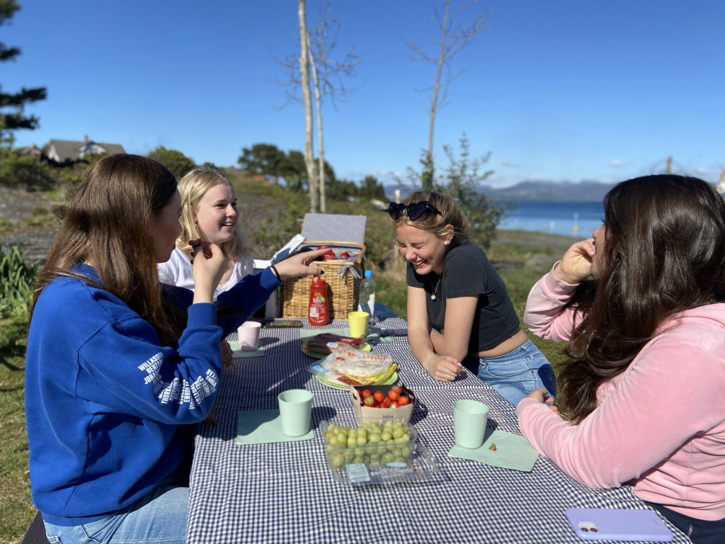 Fire jenter er på piknik sammen og spiser pølser og frukt. De ler og er glade.