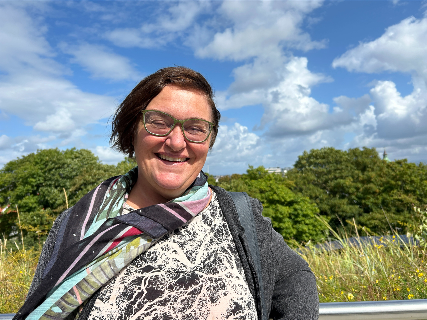 May Britt Jensen tiltrer som ny landbrukssjef i Stavanger kommune 2. september. Portrettbilde av May Britt Jensen. Hun har mørkt krøllet hår, briller og et bredt smil. I bakgrunnen er den grønne trær, og solen skinner.