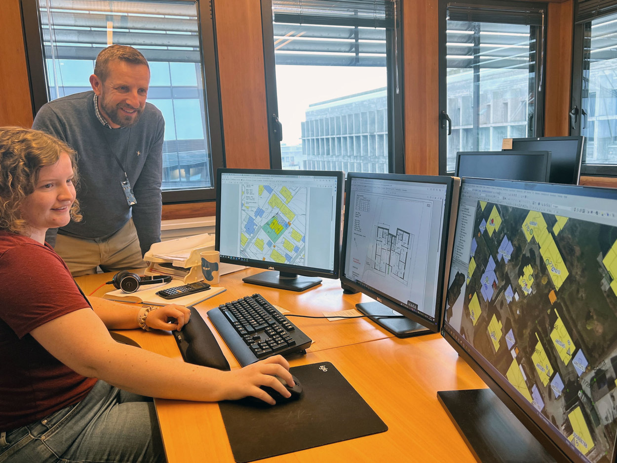 To ansatte i avdeling for kart og digitale tjenester, og PC-skjermer som viser matrikkelkart og plantegninger. Utsikt over byen gjennom vinduene. 