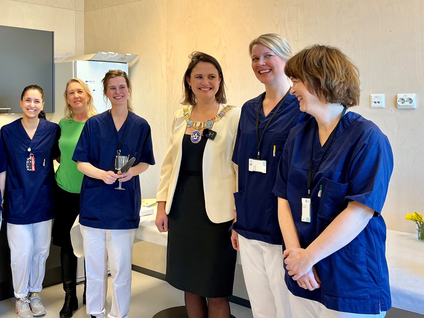 Fra høyre fastlegene Anne Karen Bøe, Kristin Eide og Stine Lise Kvinnsland, som feiret åpningen av sin nye arbeidsplass sammen med ordfører Kari Nessa Nordtun. Åse Straumstøyl, leder for de kommunale legetjenestene og Sarah Helene Hammersland, sykepleier ved legesenteret til venstre i bildet. På bildet ser vi ordfører Kari Nessa Nordtun som smiler og snakker med fem kvinner. De er leger og helsepersonell, og er kledd i blå uniformer.