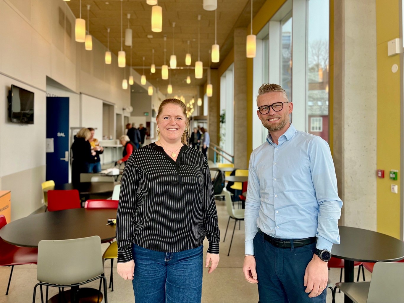 Elin Aase, rektor for Stavanger kulturskole og Helge Dragsund, kommunalsjef i helse og velferd, er storfornøyde med oppstarten av demenskoret. – Jeg brenner personlig for sang, og har sett hvordan sang kan skape ro og glede hos personer med demens, sier Aase. Vi ser Elin Aase og Helge Dragsund som står i foajeen i kulturskolen. Et stort luftig rom, med hengende lamper og bord i bakgrunnen.