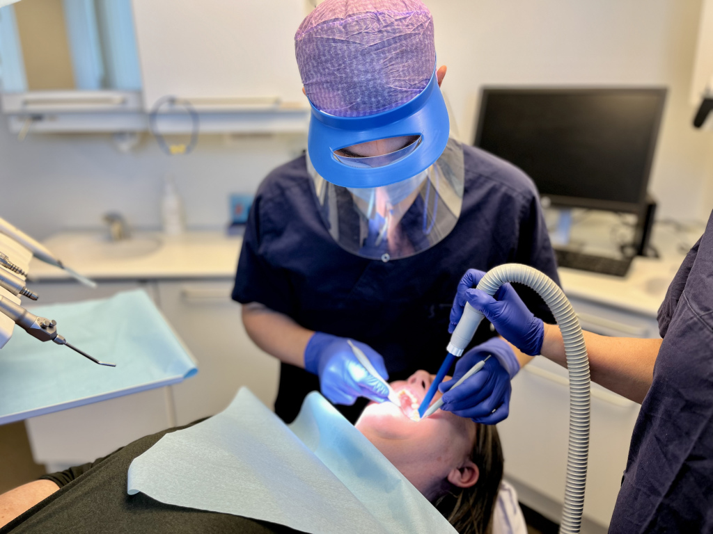En som sitter i tannlegestolen og gaper får undersøkt tennene sine av en tannlege med blått visir.