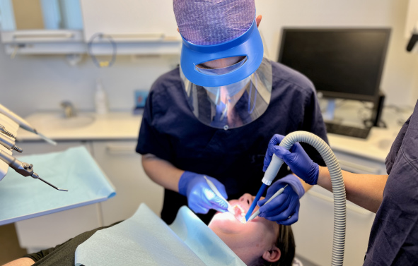 En som sitter i tannlegestolen og gaper får undersøkt tennene sine av en tannlege med blått visir.