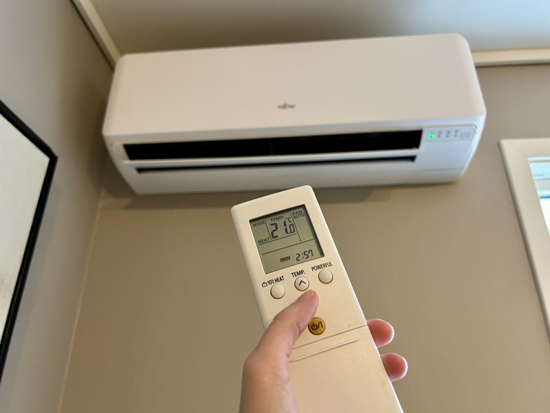 En hånd som holder en hvit fjernkontroll mot en hvit varmepumpe som henger på veggen. Temperaturen blir satt ned til 21 grader.