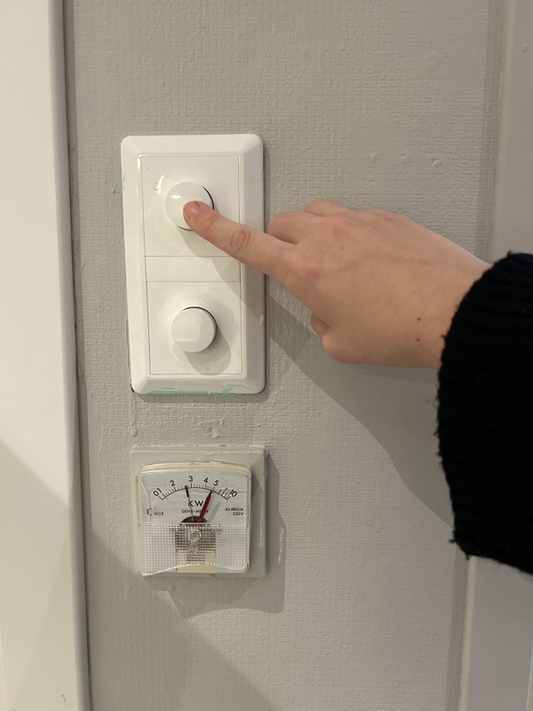 En hånd som trykker på en rund knapp på en veggmontert bryter, med en strømmåler (kW-måler) plassert under.