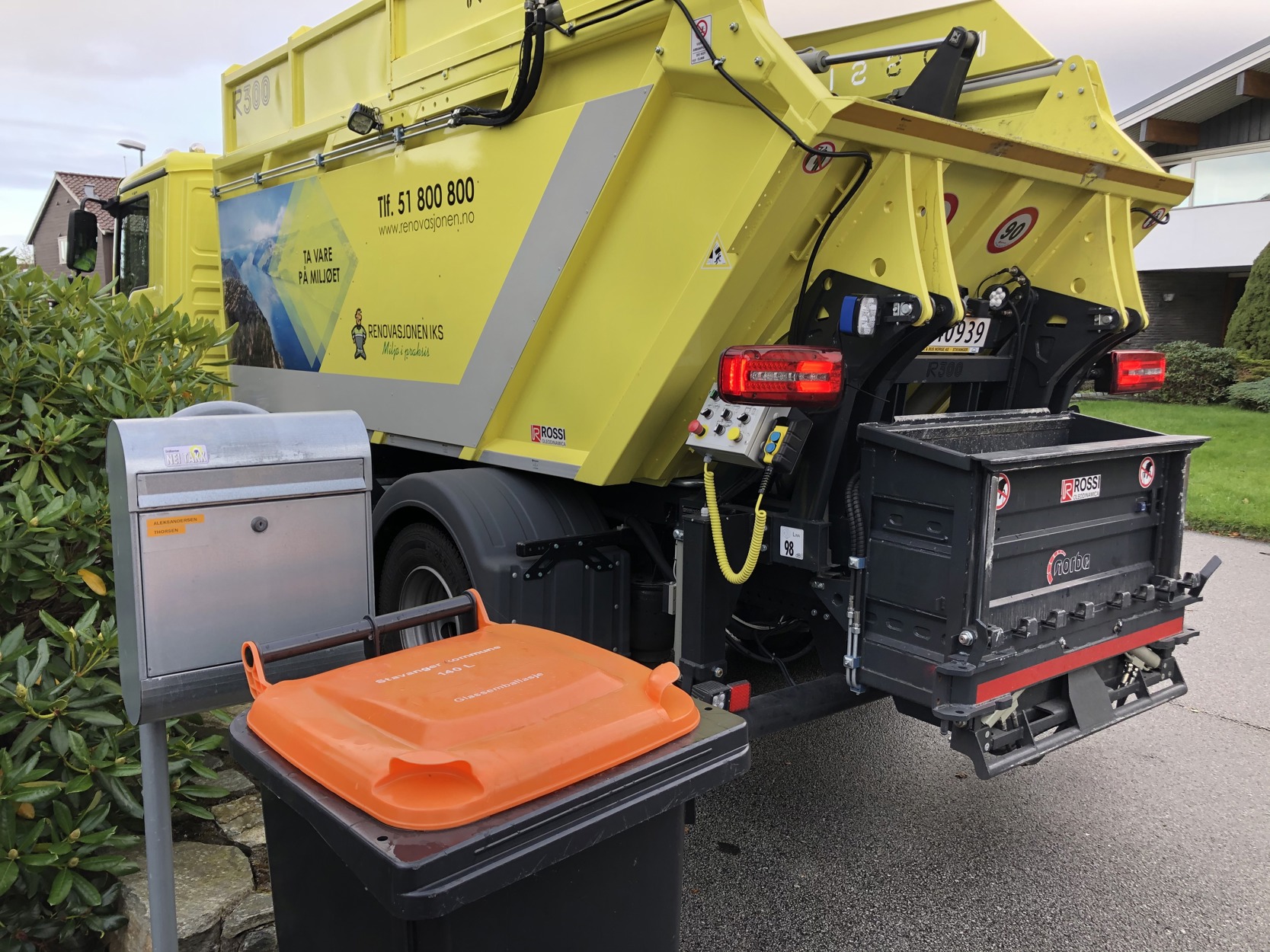 En gul søppelbil fra Renovasjonen IKS står parkert ved veikanten for å tømme avfall. Foran bilen står en søppelkasse med oransje lokk.