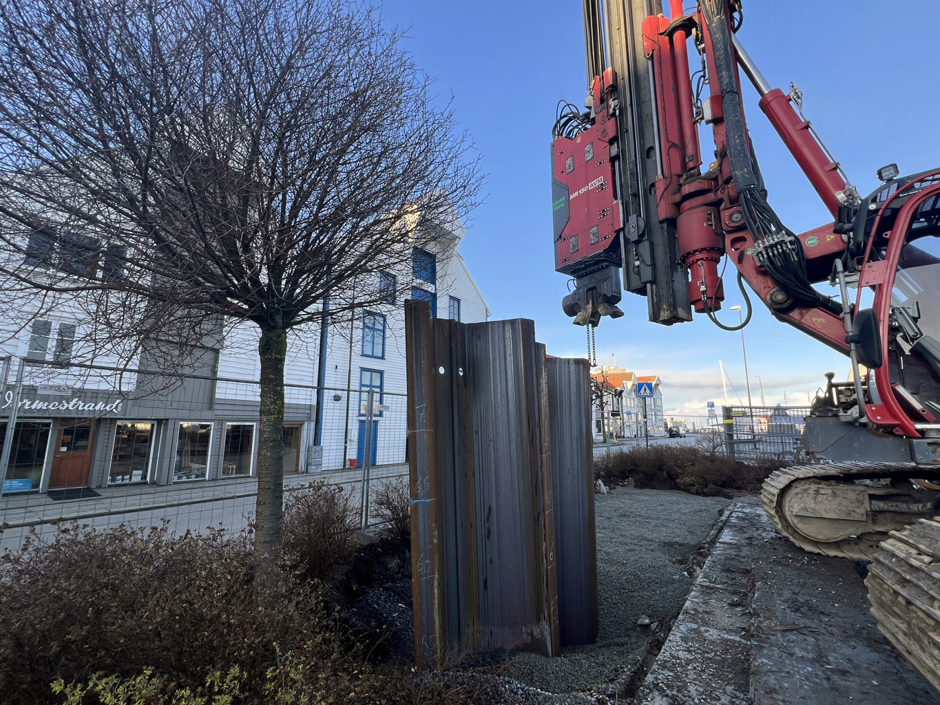 Det planlagte flomvernet består av stålplater som slås 4–6 meter ned i bakken og danner en tett barriere under jorden. Den ferdige veggen vil stikke 50–80 cm opp over grunnen og beskytter mot stormflo i framtiden. Flomvernet kan integreres i bylandskapet som en voll, eller ved hjelp av urbane møbler. Bildet er fra prøvespuntingen ved Kjerringholmen i november 2024. Flere meter høye stålplater slås ned i grunnen ved kjerringholmen. (Foto: Sigrun Sætrevik)