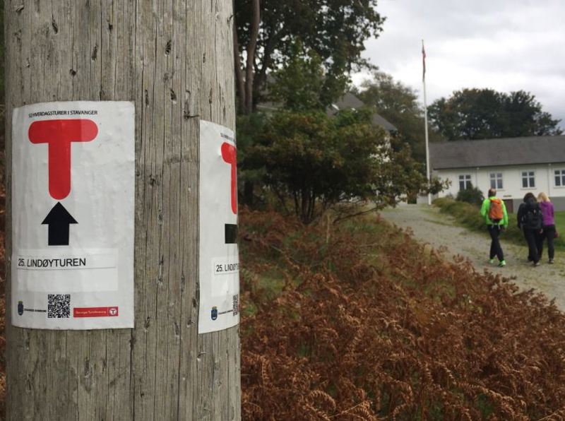 Stolpe med skilt for Lindøyturen, ved Lindøy kai. I bakgrunnen ett av de hvite husene ved kaien.