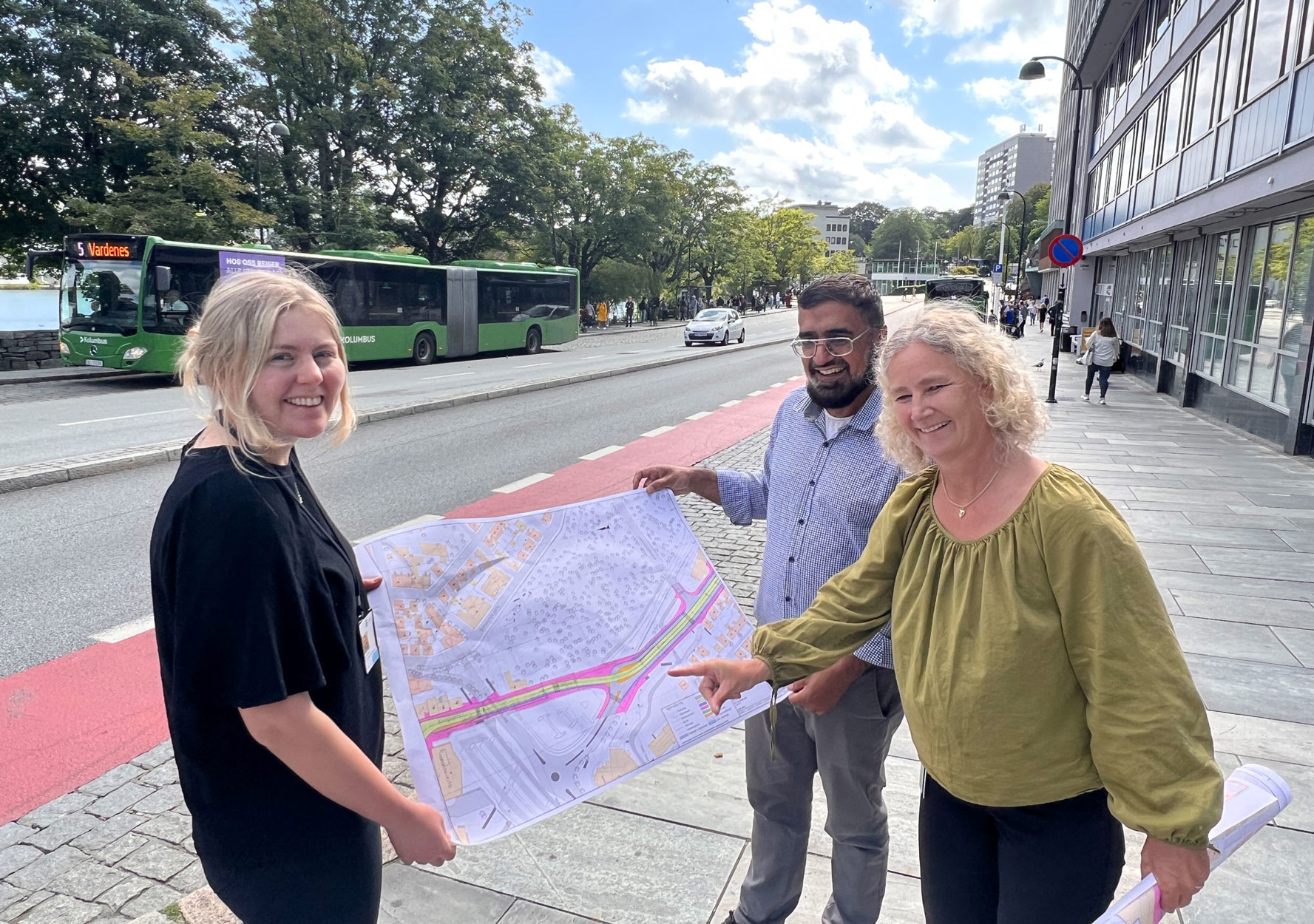 Tre transportplanleggere holder opp et plankart ute i gata, med buss og sykkelfelt i bakgrunnen.