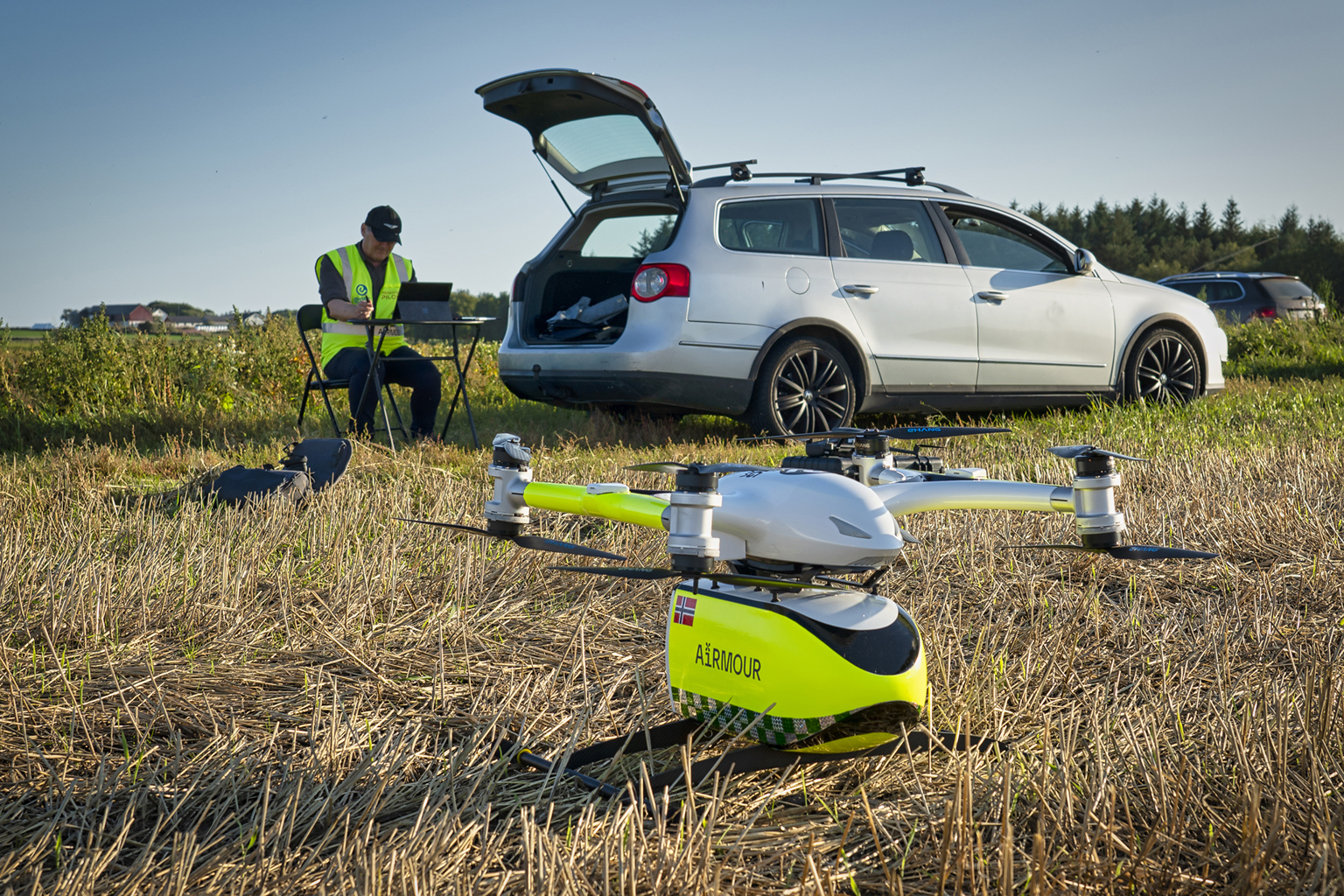 Foto: eHang Scandinavia. En mann med refleksvest og kaps sitter med en pc på en eng. Ved siden av står en lysgrå bil med bakluken åpen. Noen meter foran mannen og bilen står en hvit og refleksgul drone klar til å fly, med en gul beholder under hvor det står Airmour skrevet på siden, sammen med et lite norsk flagg.