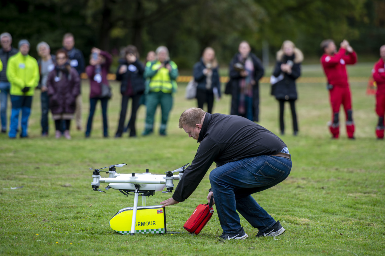 En mann henter ut en rød hjertestarter fra det gule lasterommet på en hvit drone som står på bakken. I bakgrunnen står det flere mennesker og følger med.