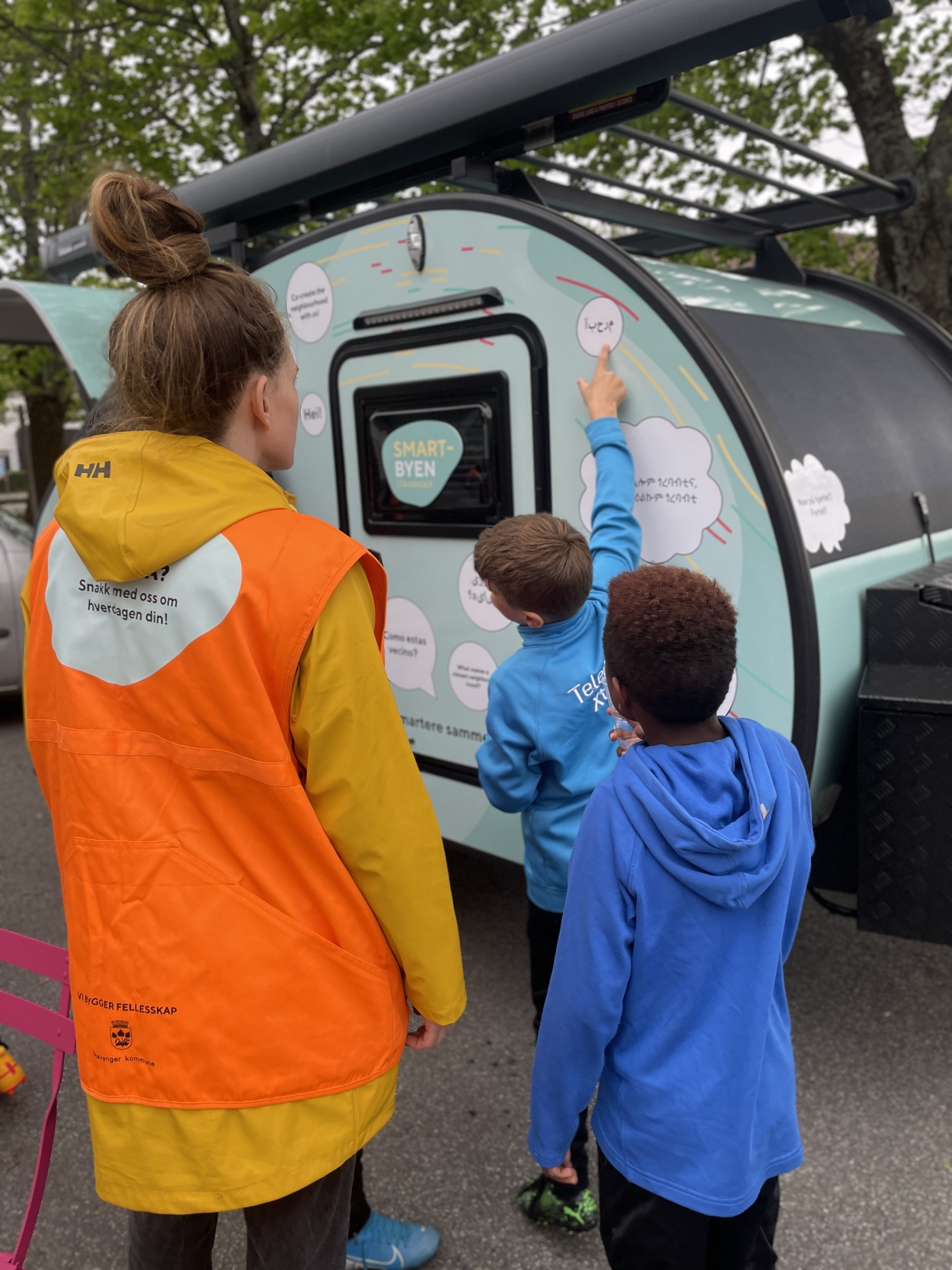 En dame og to gutter ser på gampingvogna. Gutten peker på en snakkeboble der det står hei på polsk.
