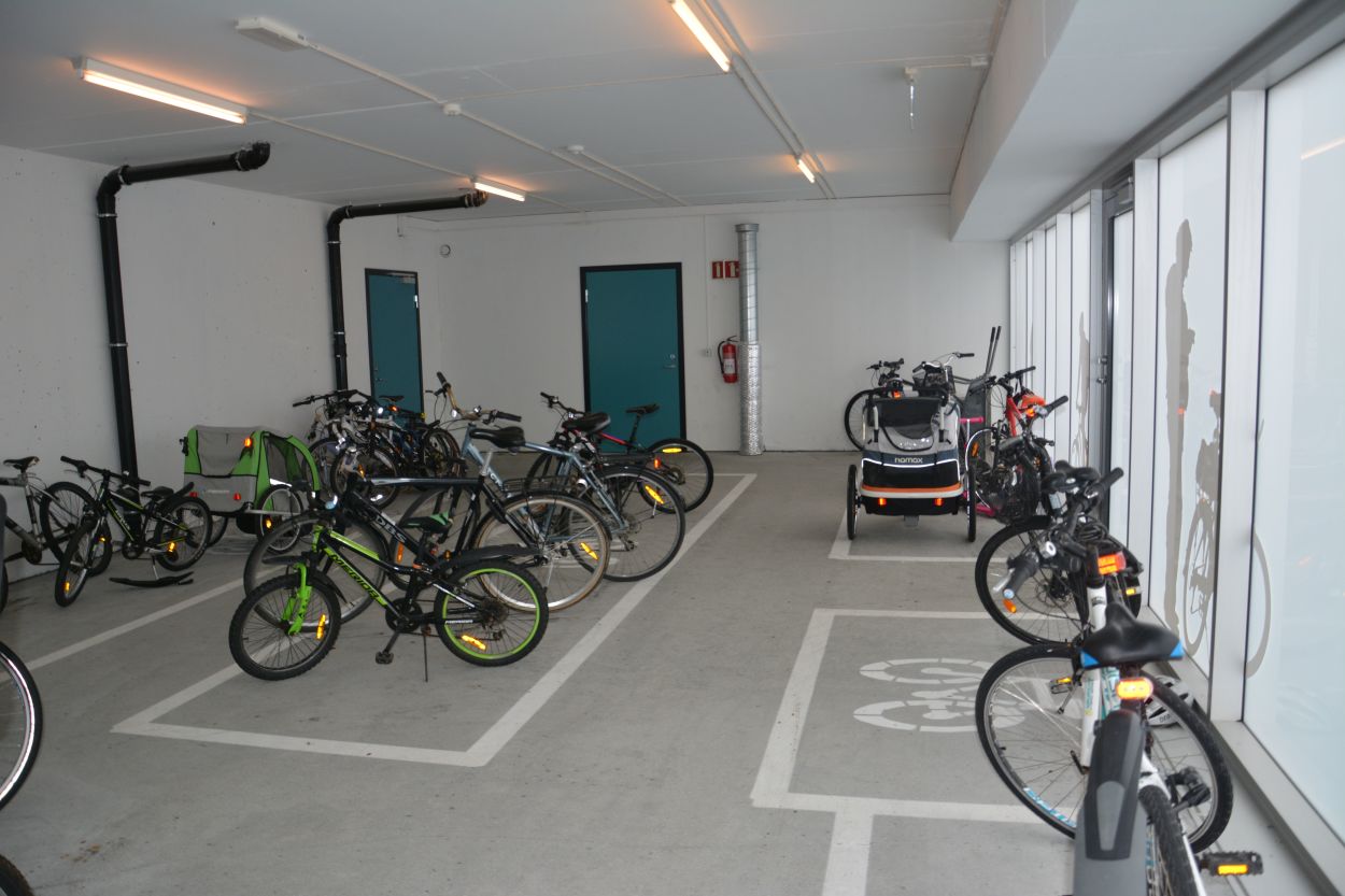 I et stort rom står barn- og voksensykler og sykkelvogn parkert. På gulvet er det oppmerket hvite linjer og sykkelsymboler slik at det blir tydelig hvor syklene skal parkeres i rommet.