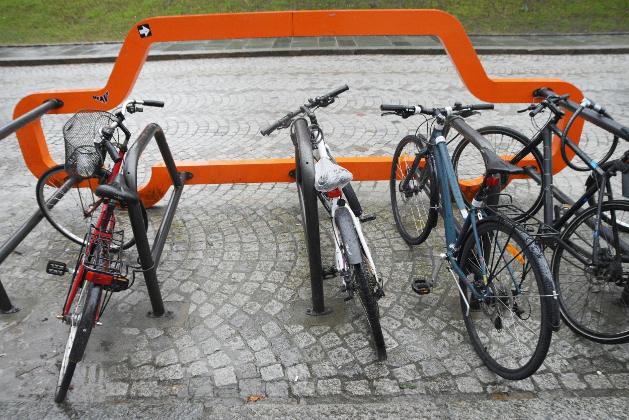 Bak en oransje skulptur som likner en bil er det 5 sykkelstativer montert. Fire sykler er låst ved stativene.