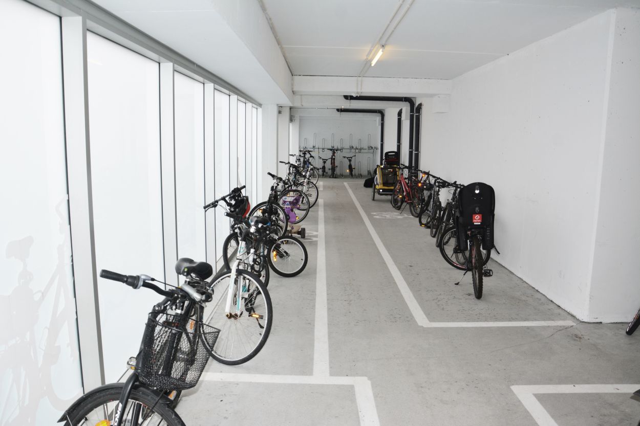 I ett langstrekt rom står flere sykler parkert uten stativer. I bakgrunnen står et toetasjes stativ med sykler i.