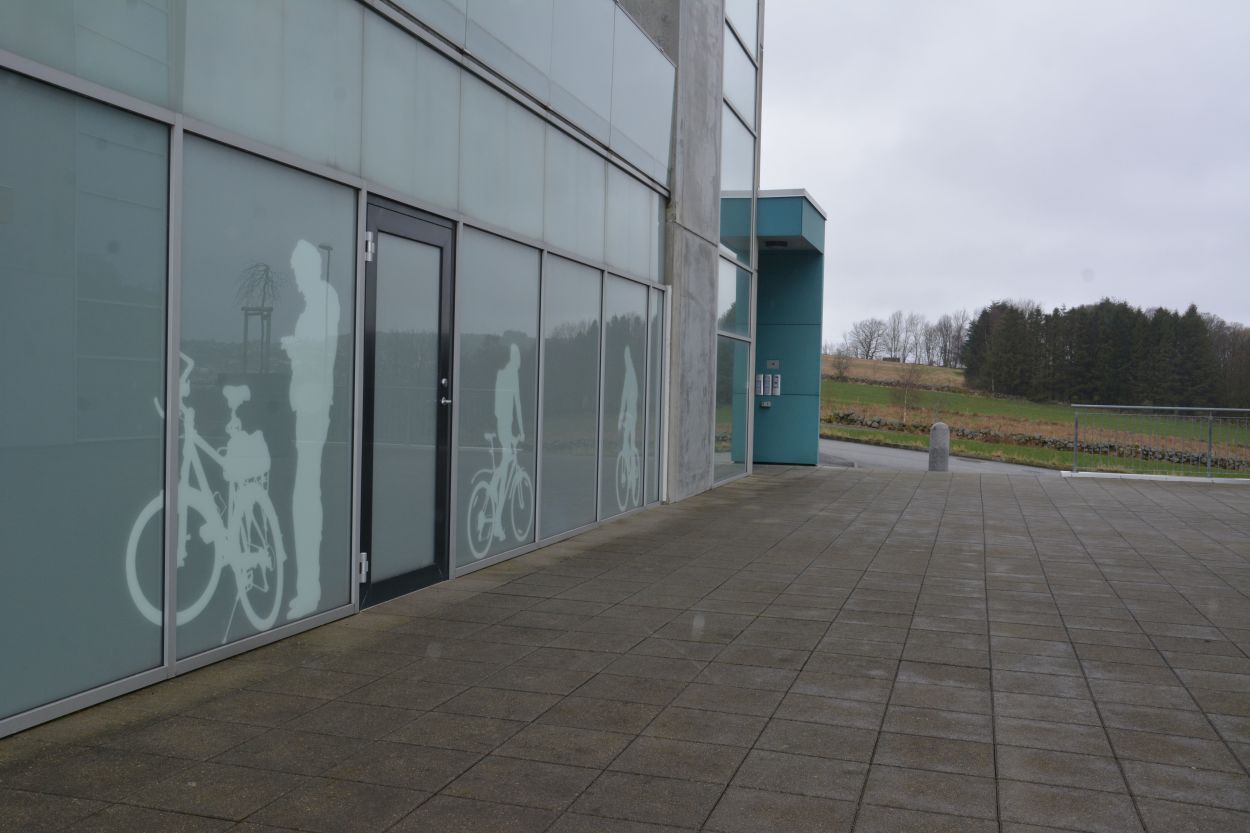 På veggene ved siden av inngangen til et innendørs sykkelparkeringsanlegg finnes det ca. to meter store illustrasjoner som viser personer med sykler.