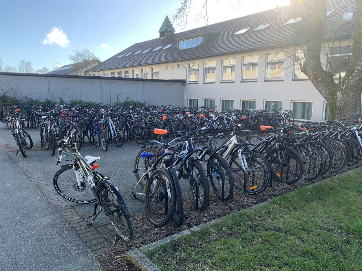 Ved siden av en skole står mange sykler parkert i stativer på en asfaltert sykkelparkeringsplass.