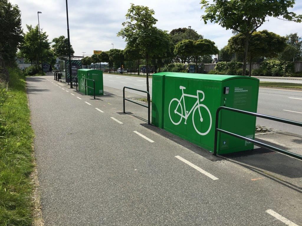 Ved siden av en bussholdeplass står to grønne bokser der man kan parkere og låse inn  sykkelen sin. På sideveggene av boksene finnes det en stor sykkelsymbol i hvit.