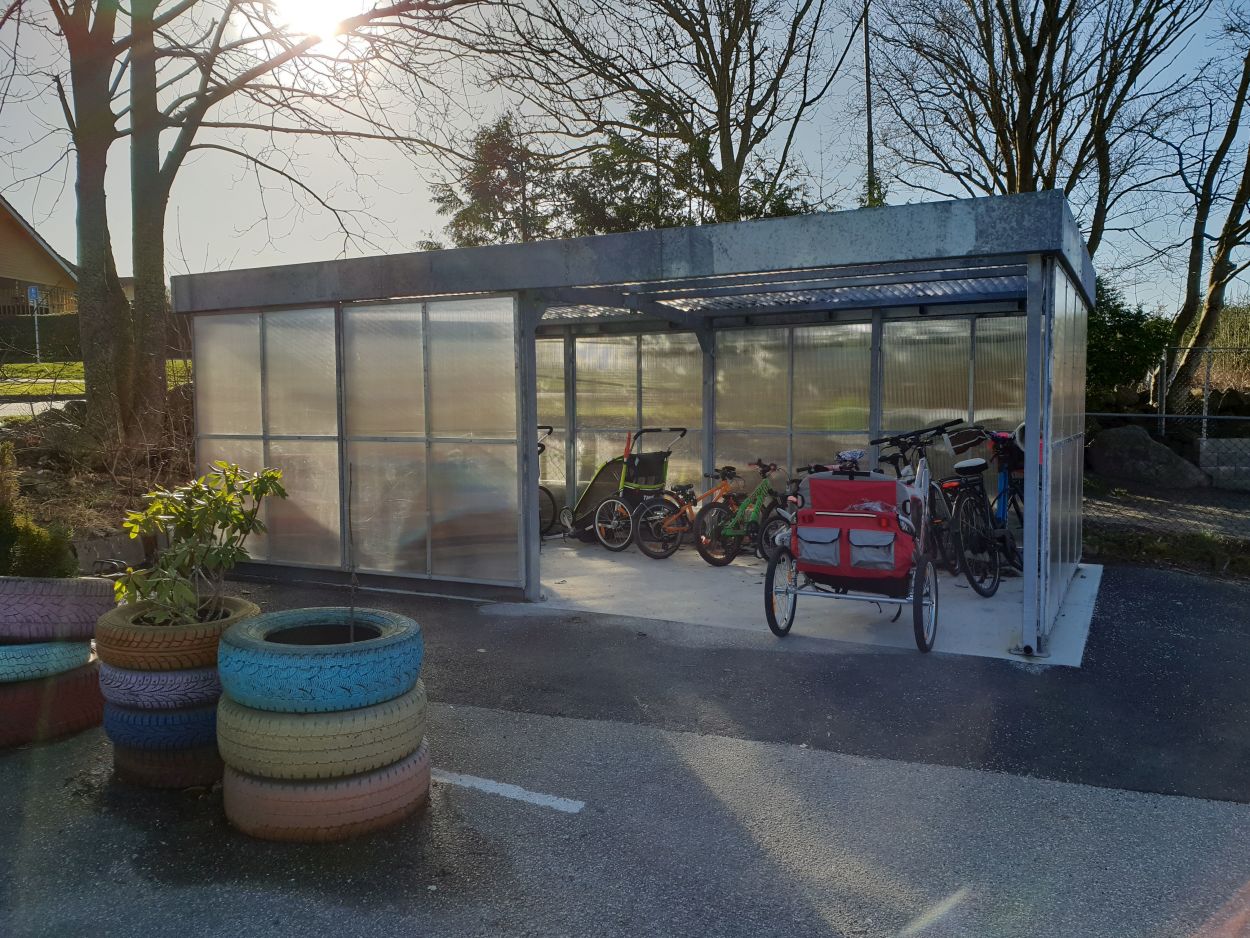 I et sykkelvognhus utenfor barnehagen med gjennomsiktige vegger står flere sykkelvogn samt voksen- o barnesykkel parkert.