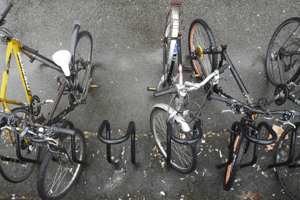 Noen sykler står parkert i noen sykkelstativer. På bakken ligger det papirposer og mange sneiper. Et sykkel mangler sete.