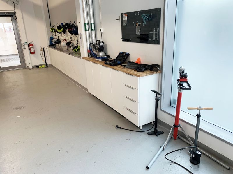 I innendørs sykkelparkering hos Statens vegvesen i Stavanger henger flere sykkelhjelm på knagger på en vegg. Ved siden av en benk med verktøy står to sykkelpumper.