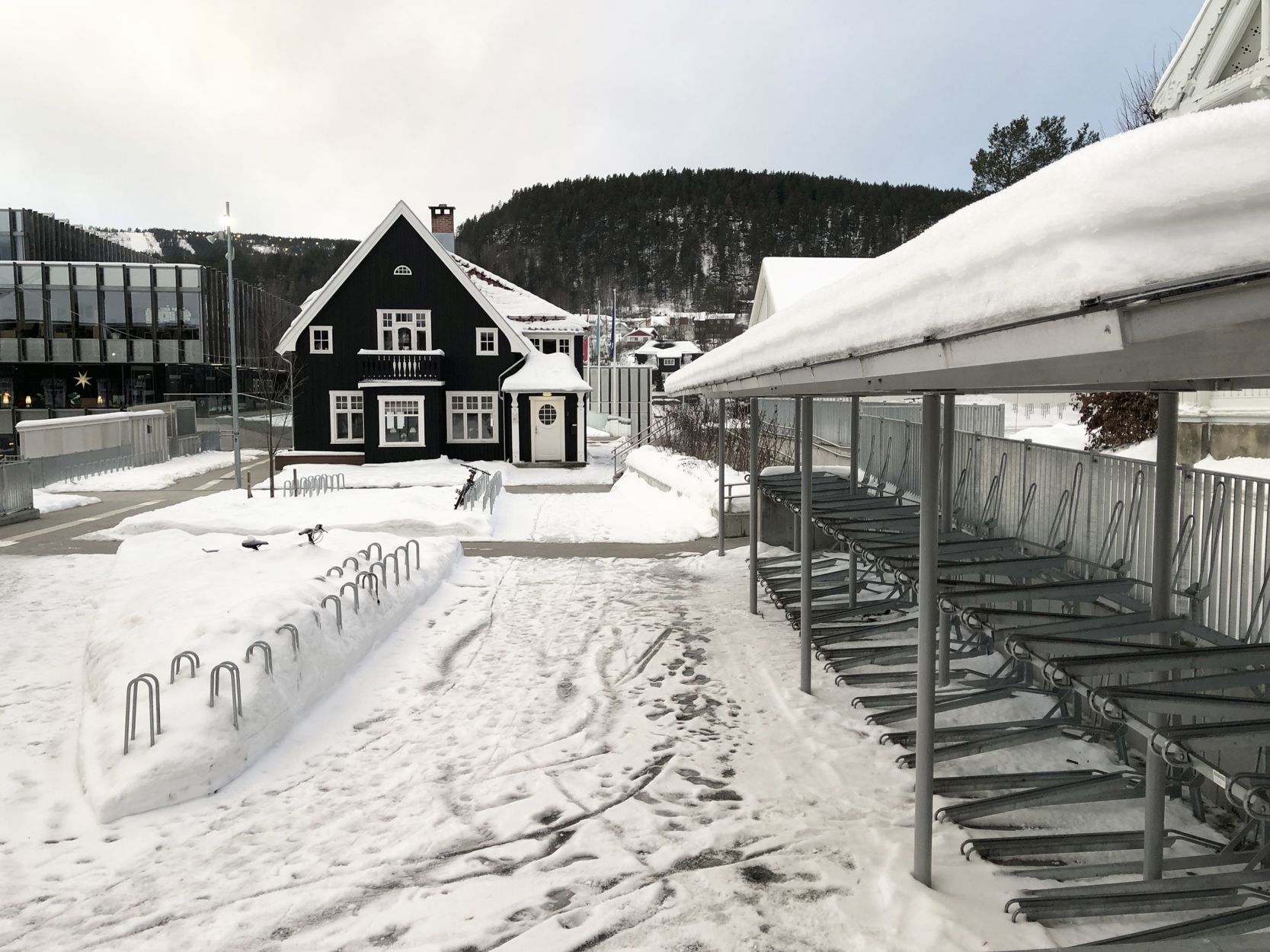 På en untendørs sykkelparkeringsplass ligger det mye snø mellom stativene uten overdekk mens de toetasjes stativer som har overdekk er tilgjengelig og kan brukes.